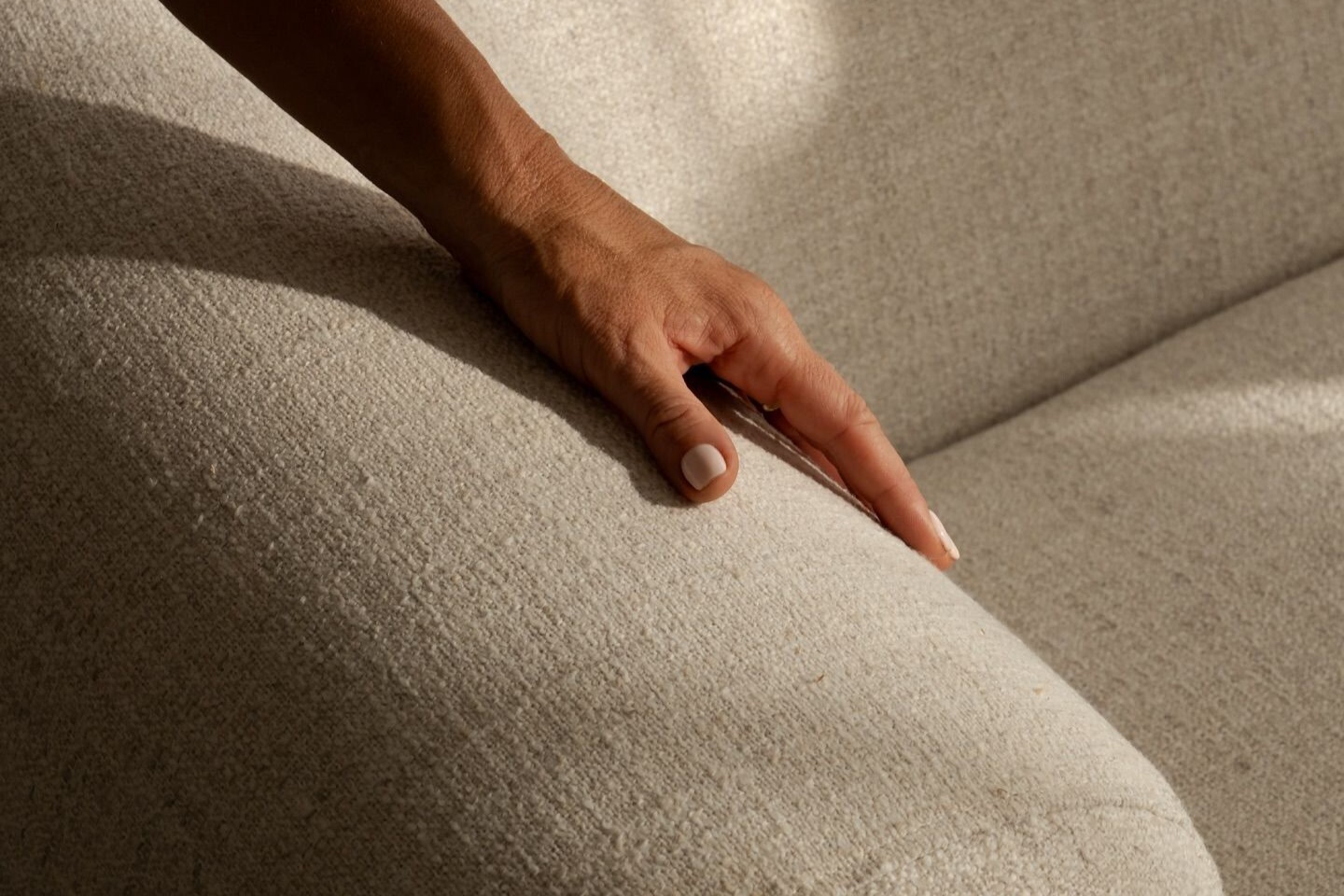 Hand mit hellem Nagellack liegt auf beigem Stoffsofa im sanften Licht.