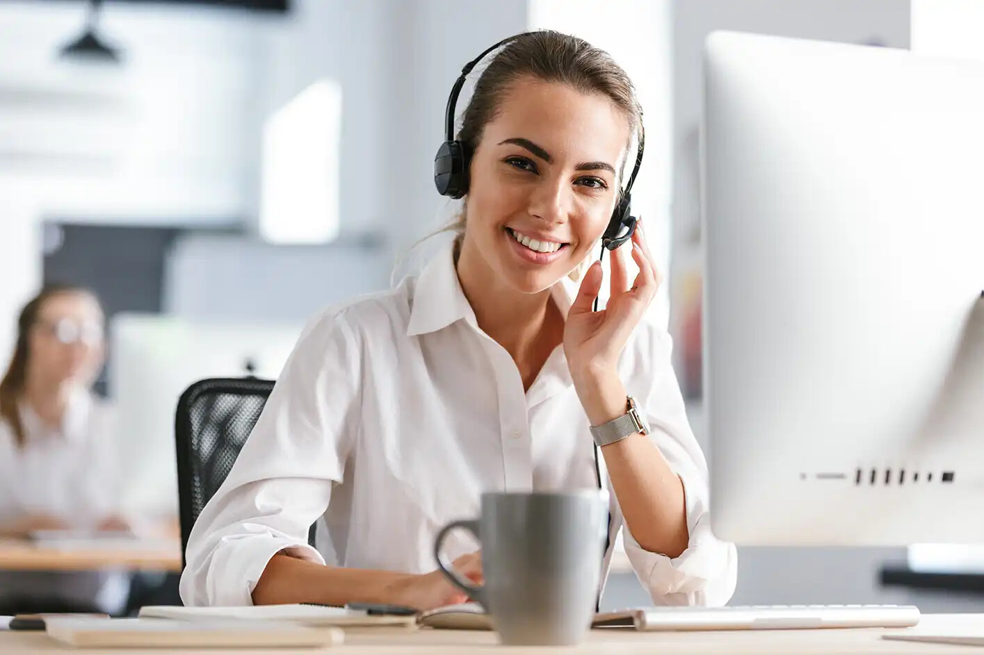 Frau mit Headset arbeitet am Computer, Kaffeetasse am Schreibtisch.