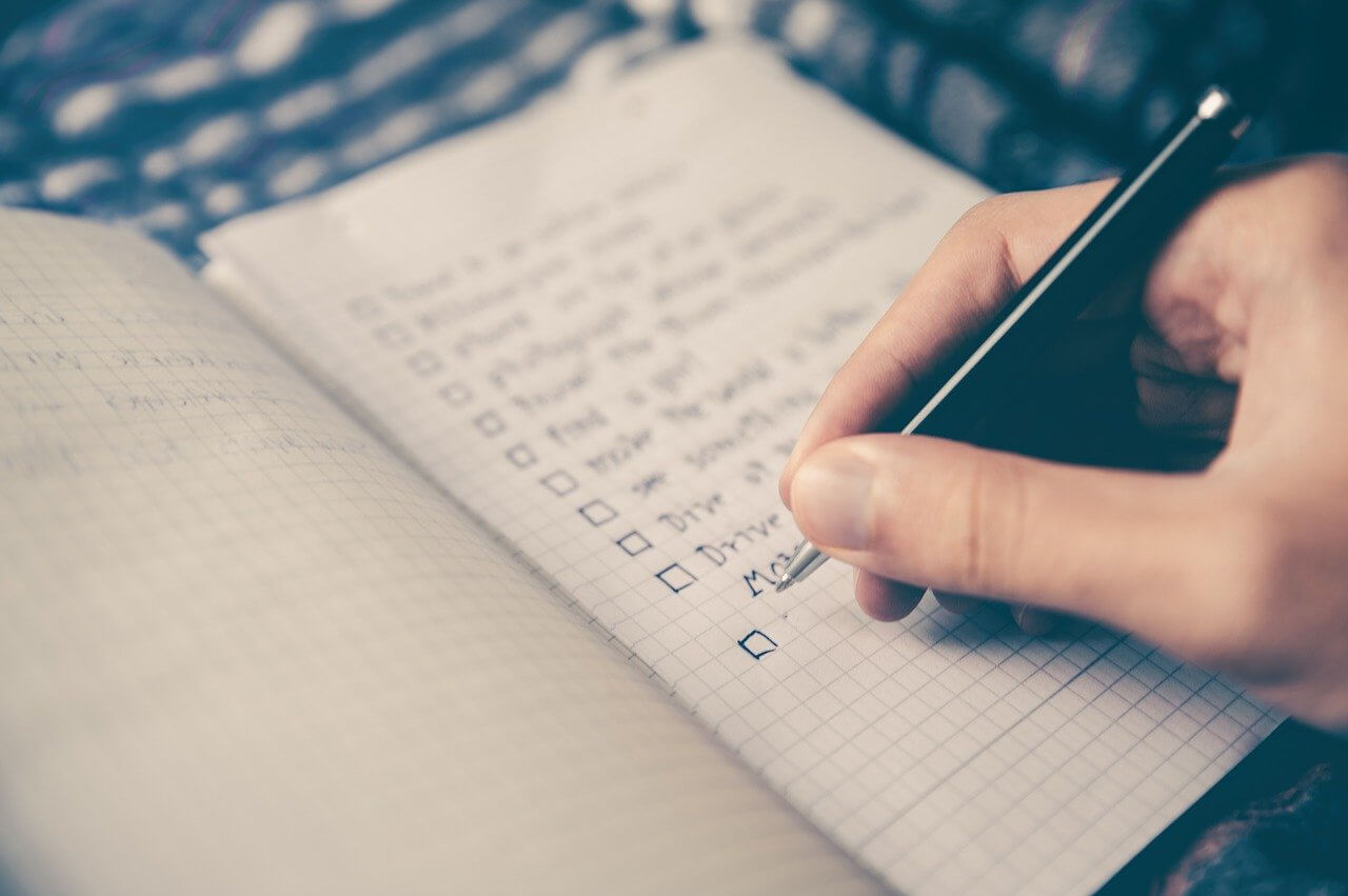 Hand schreibt Checkliste in kariertes Notizbuch auf einen Tisch.