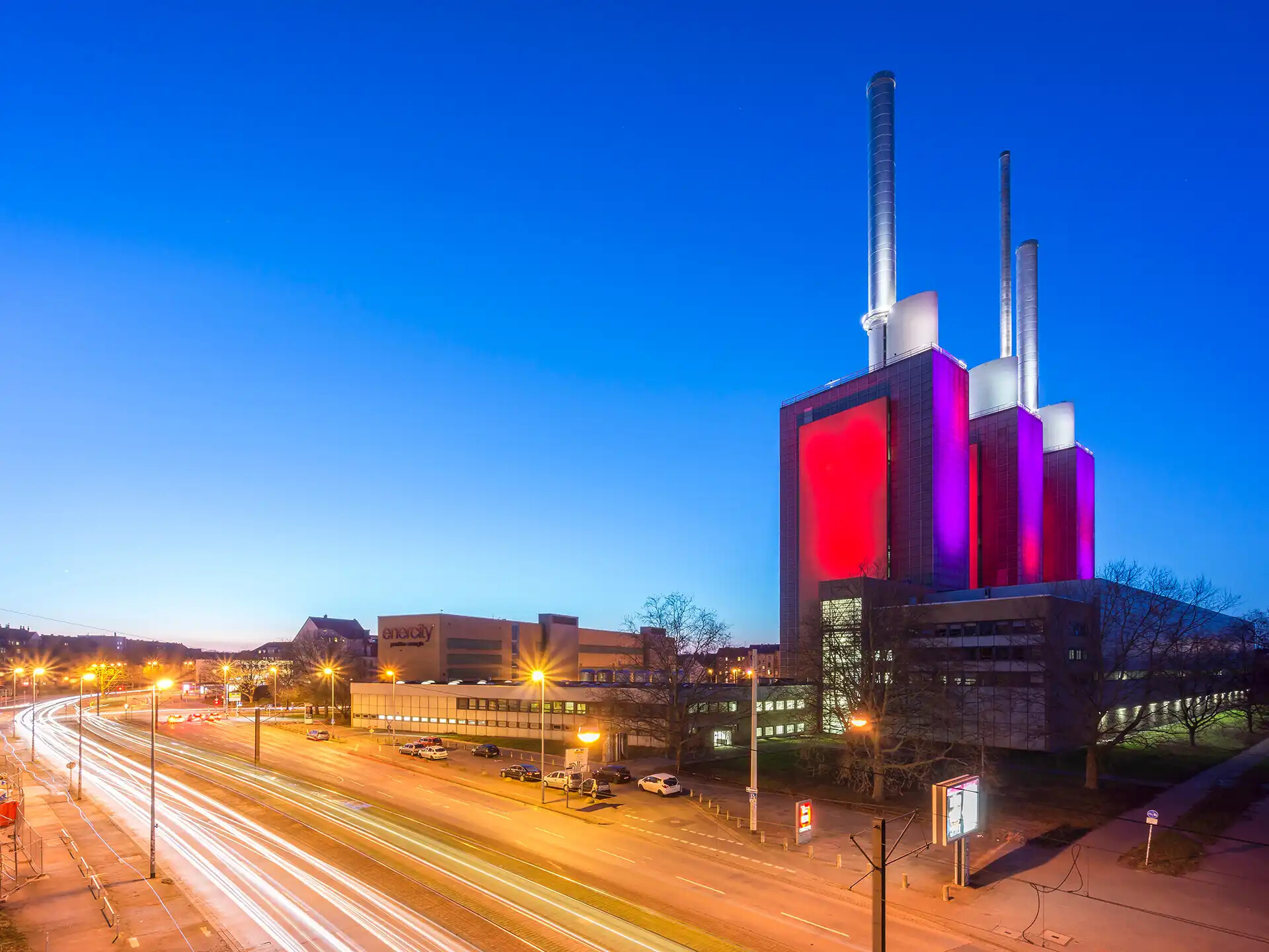 Kraftwerk mit rosafarbenen Lichtern an einer viel befahrenen Straße in der Abenddämmerung, Lichtspuren.