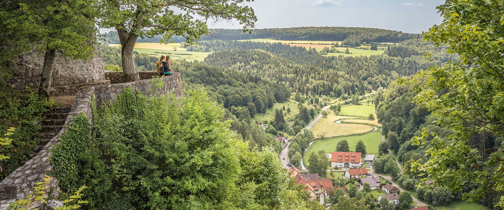 Blick von einem bewaldeten Hügel auf ein Tal mit Feldern, Wäldern und einem Dorf im Hintergrund