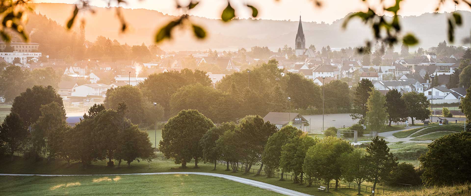 Eine Stadt im Tal, beleuchtet von warmem Sonnenlicht, mit einem prominenten Kirchturm, umgeben von grünen Feldern und sanften Hügeln.