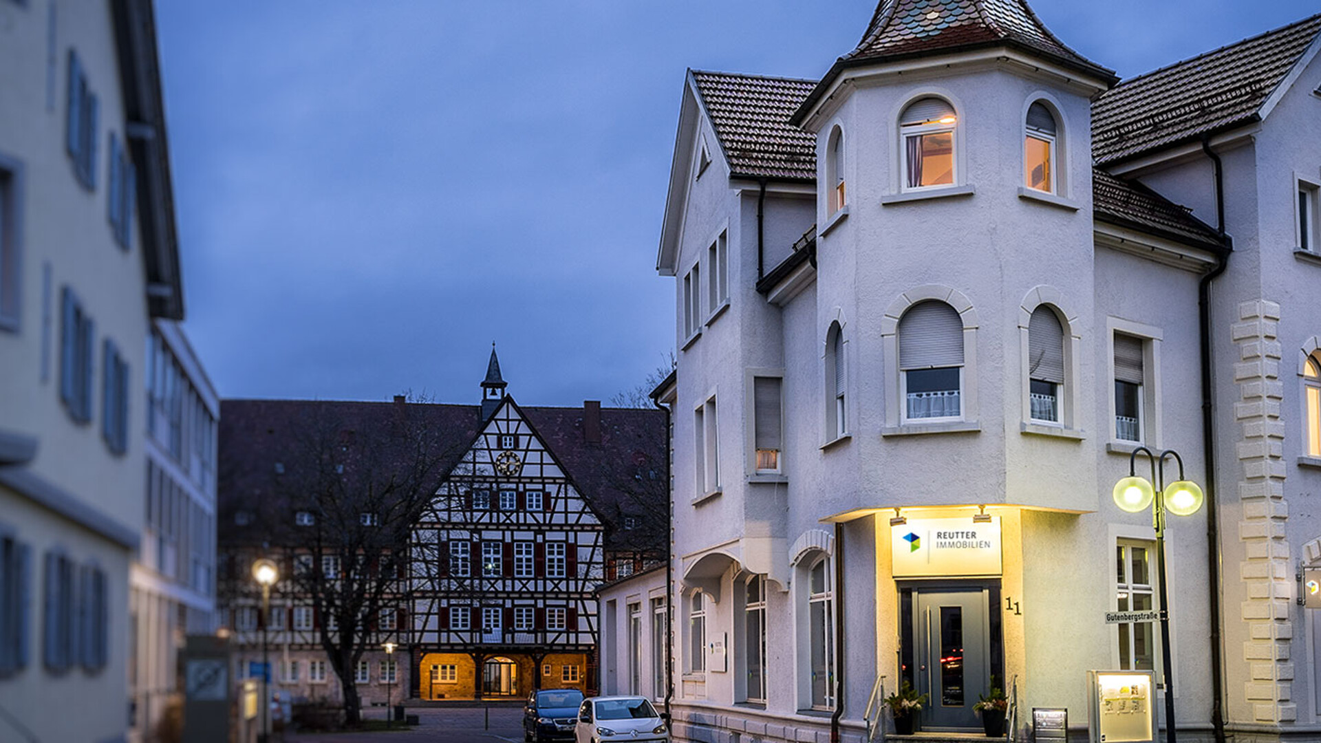 Beleuchtete Fachwerkhäuser und moderne Gebäude an einer ruhigen Straße bei Dämmerung