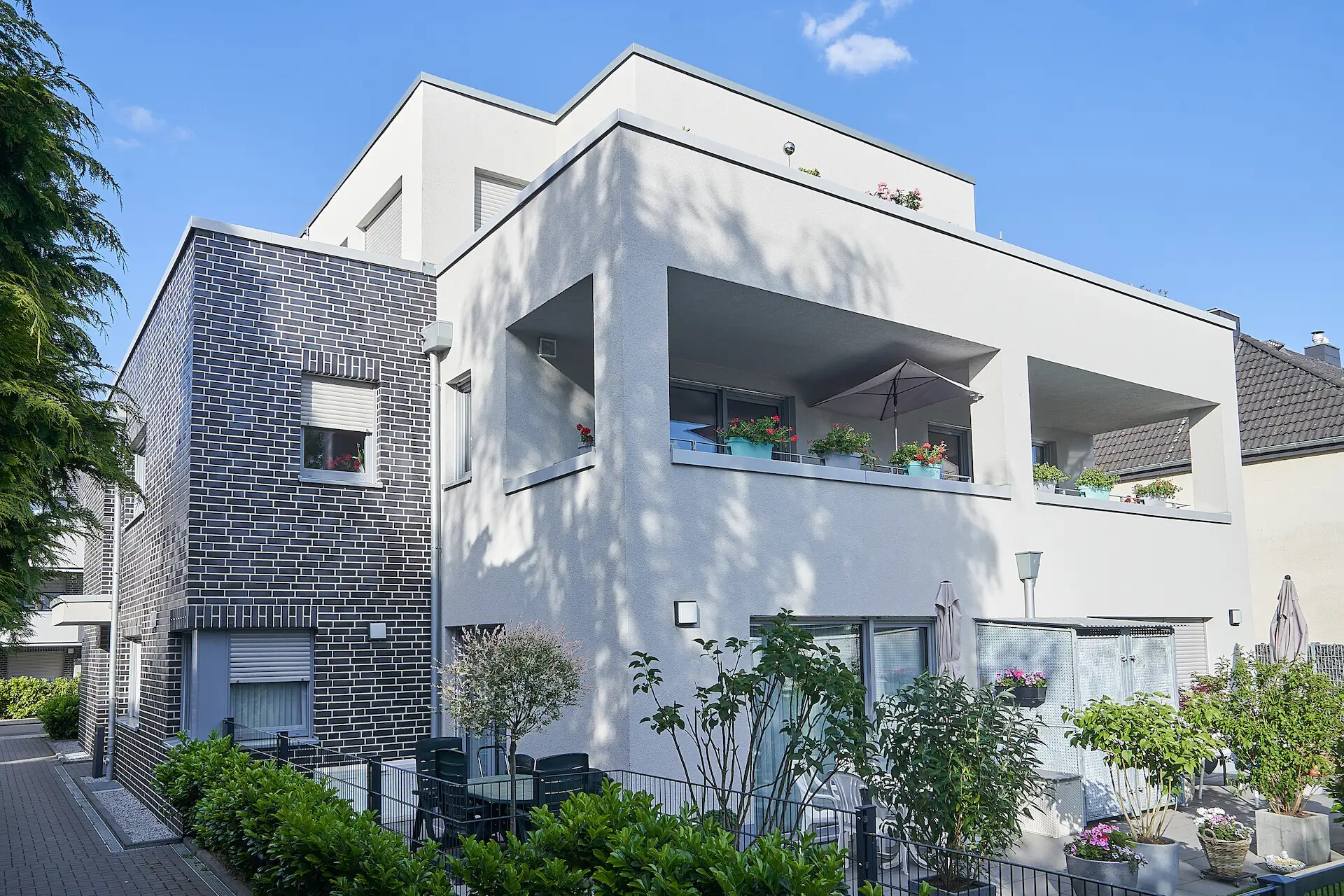 Weißes Gebäude, große Balkone, Pflanzen und Sitzgelegenheiten im Freien in der Sonne.