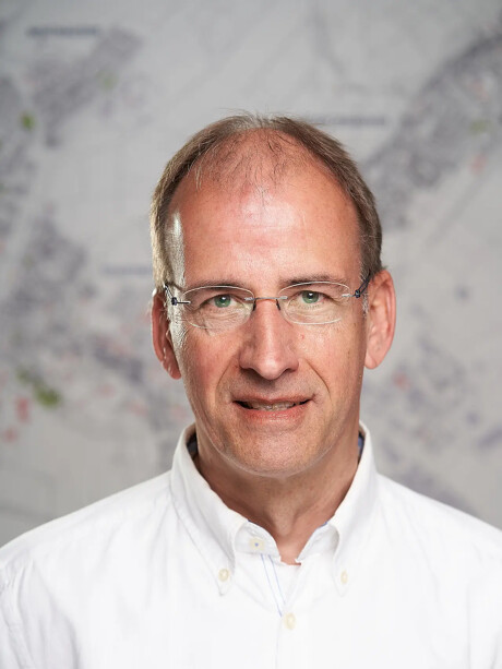Uwe Wiegand mit Brille und weißem Hemd vor unscharfer Kartenkulisse.