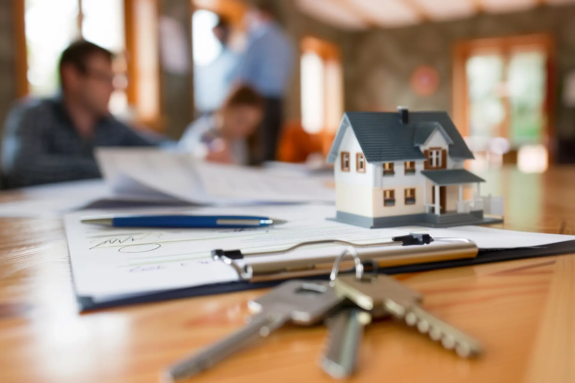 Schlüssel, Dokumente und Spielzeughaus auf Tisch, Menschen im Gespräch.