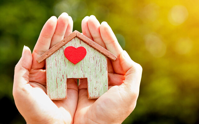 Frauenhände halten ein Holzhaus mit Herz: Das Eigenheim verkaufen