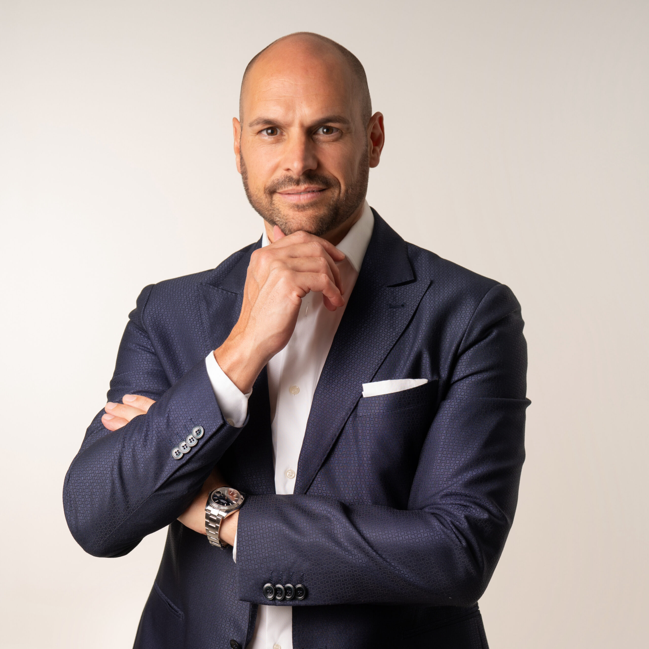 Ein glatzköpfiger Mann im dunklen Anzug posiert mit der Hand am Kinn vor einem schlichten Hintergrund.