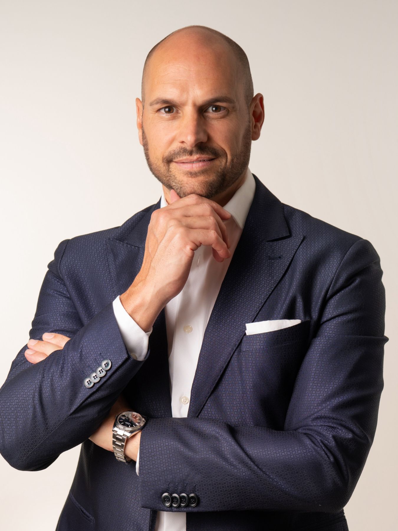 Uomo calvo in abito blu posa con la mano sul mento, sfondo semplice.