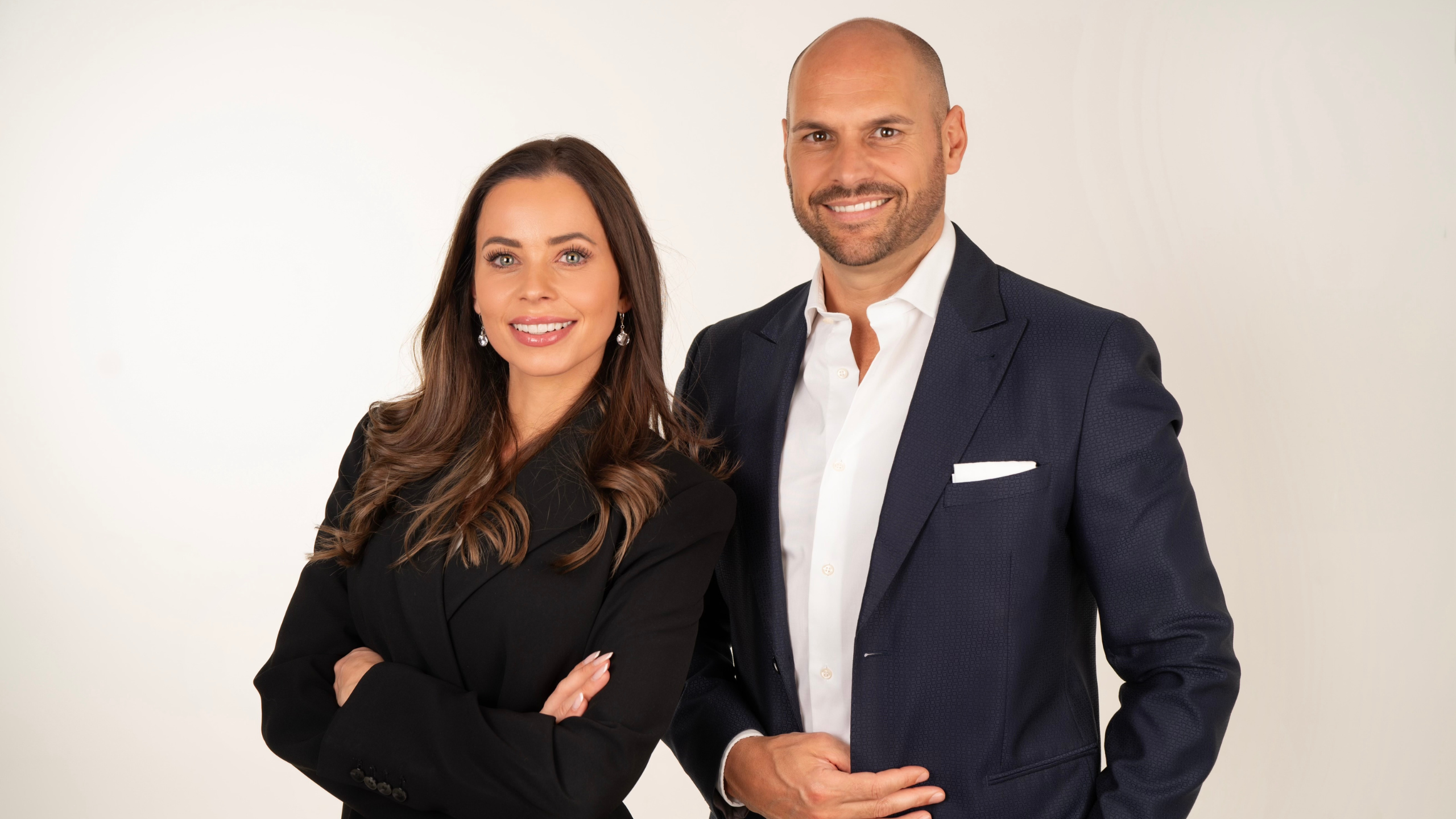 Frau und Mann, schick gekleidet, lächeln vor neutralem Hintergrund.