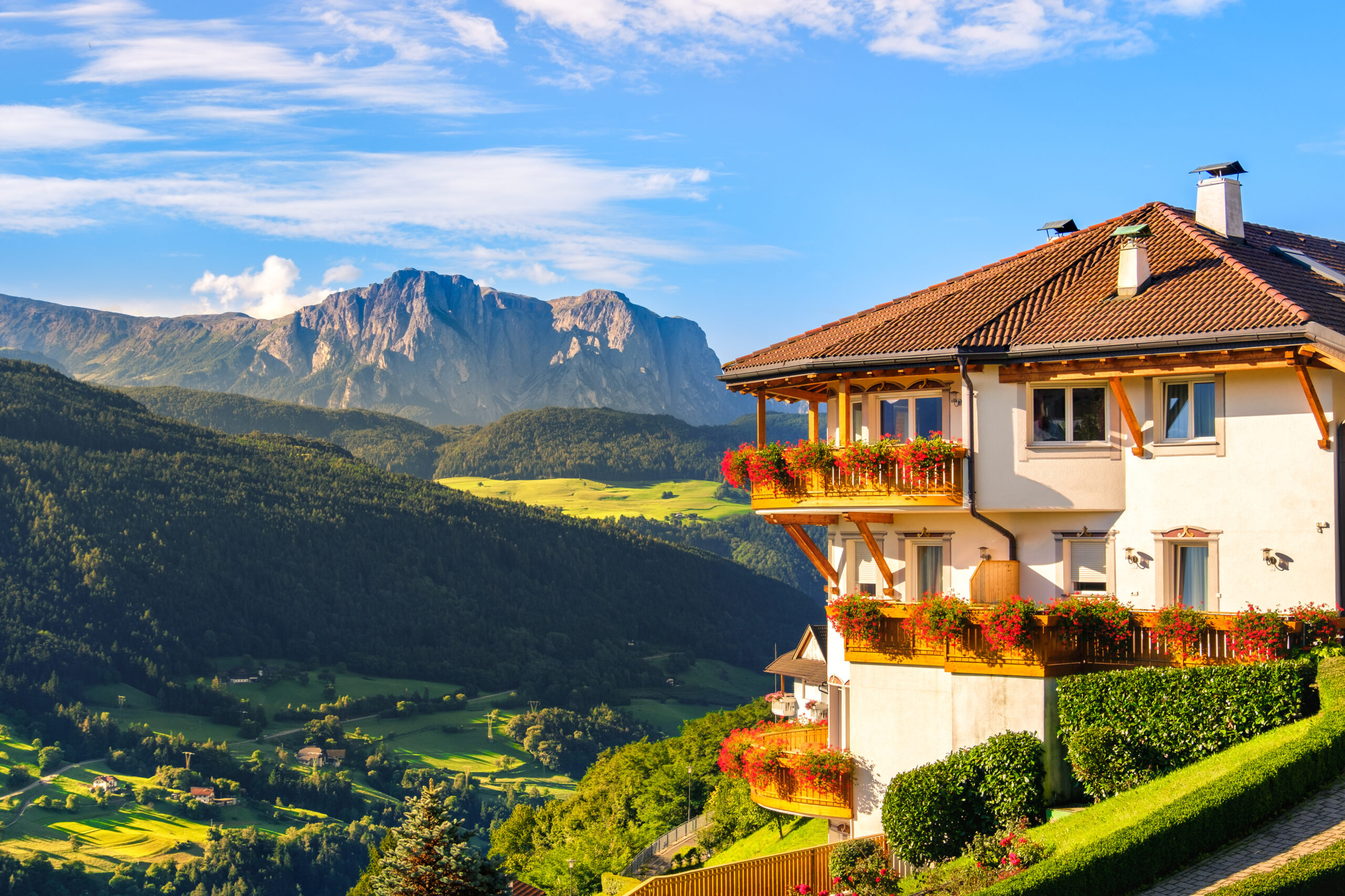 House with flowered balconies, green hills, and mountains beyond.