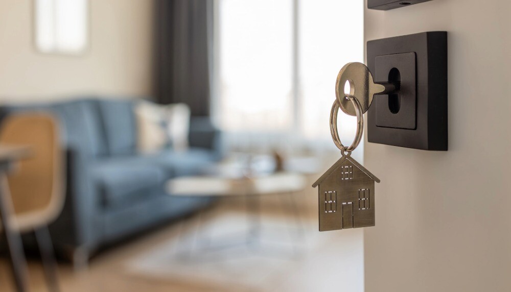 Hausförmiger Schlüsselanhänger an einem Schlüssel in einer Tür, dahinter ein verschwommener Raum.