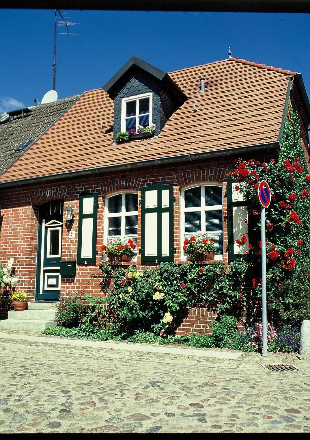 Backsteinhaus mit Blumen, grünen Fensterläden, Parkverbotsschild.