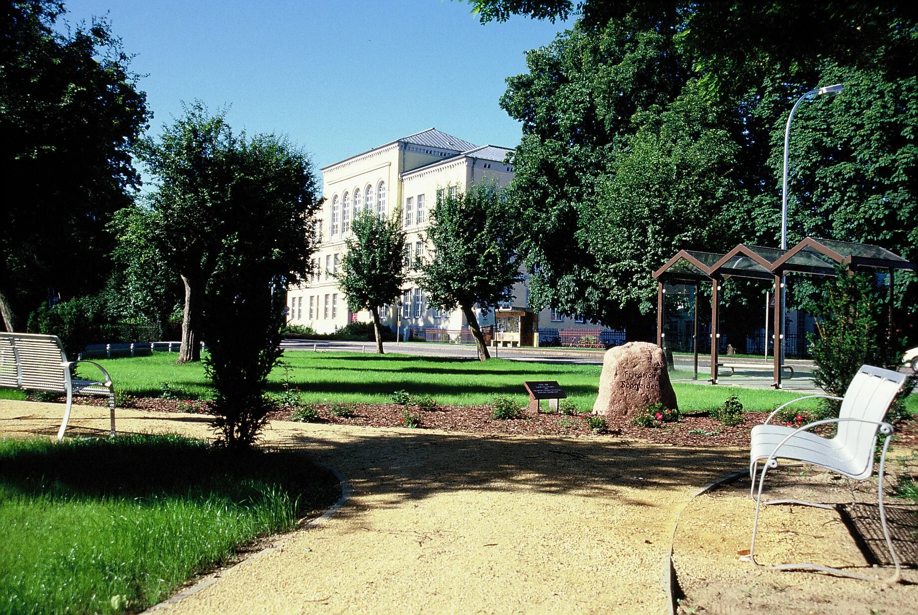Sonniger Park mit Bänken, Bäumen und weißem Gebäude.