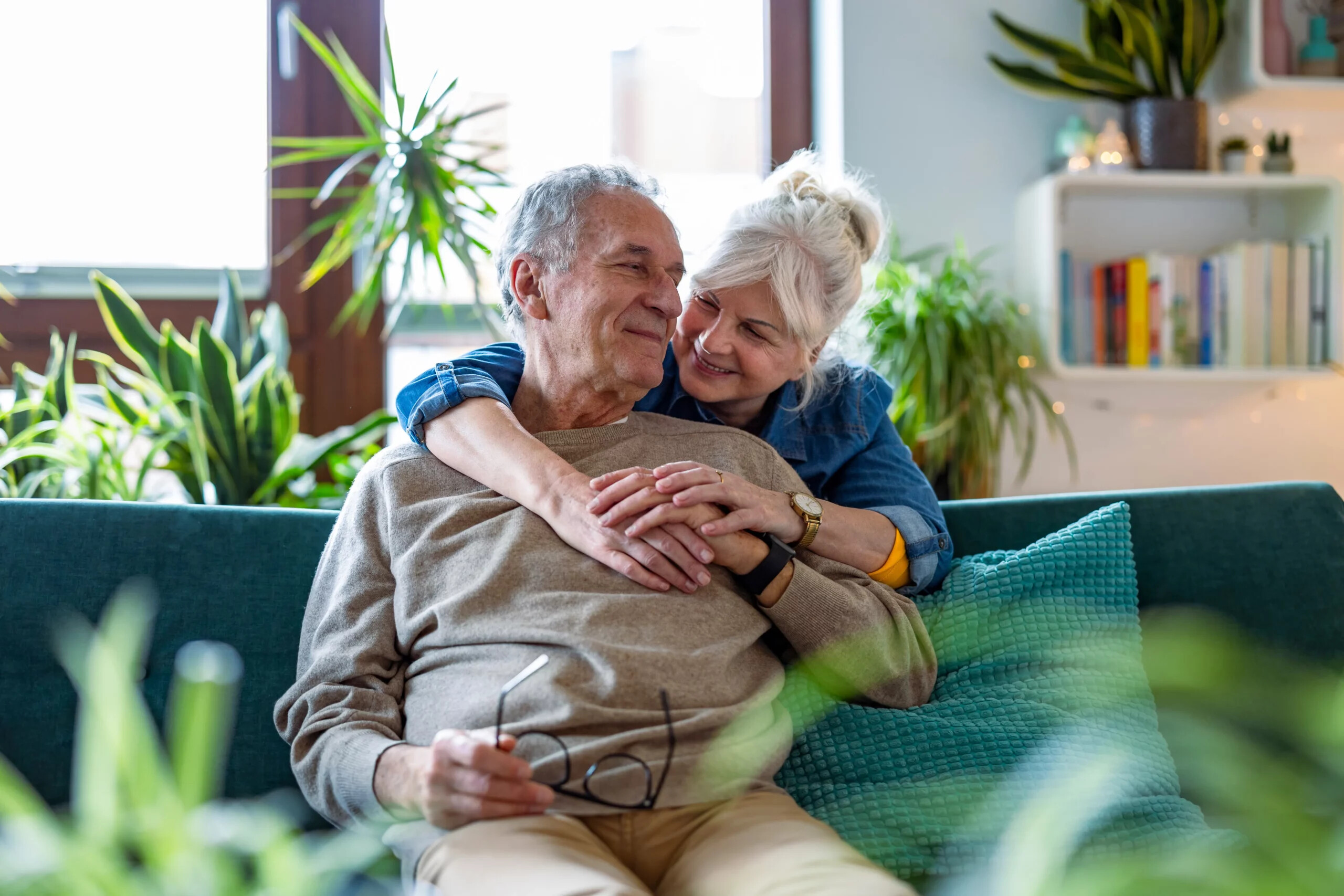 Eine Frau umarmt ihren Partner von hinten, dieser sitzt auf einer Couch