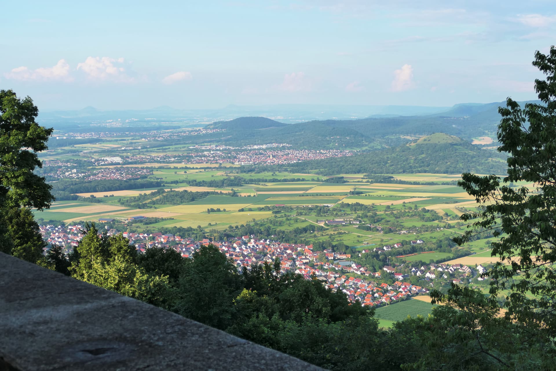 Eine Landschaft mit Feldern, Häusern und Hügeln unter blauem Himmel, Bäume.