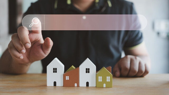 Mann klickt auf ein Browser-Suchfeld für die Immobiliensuche, im Hintergrund Modellhäuser
