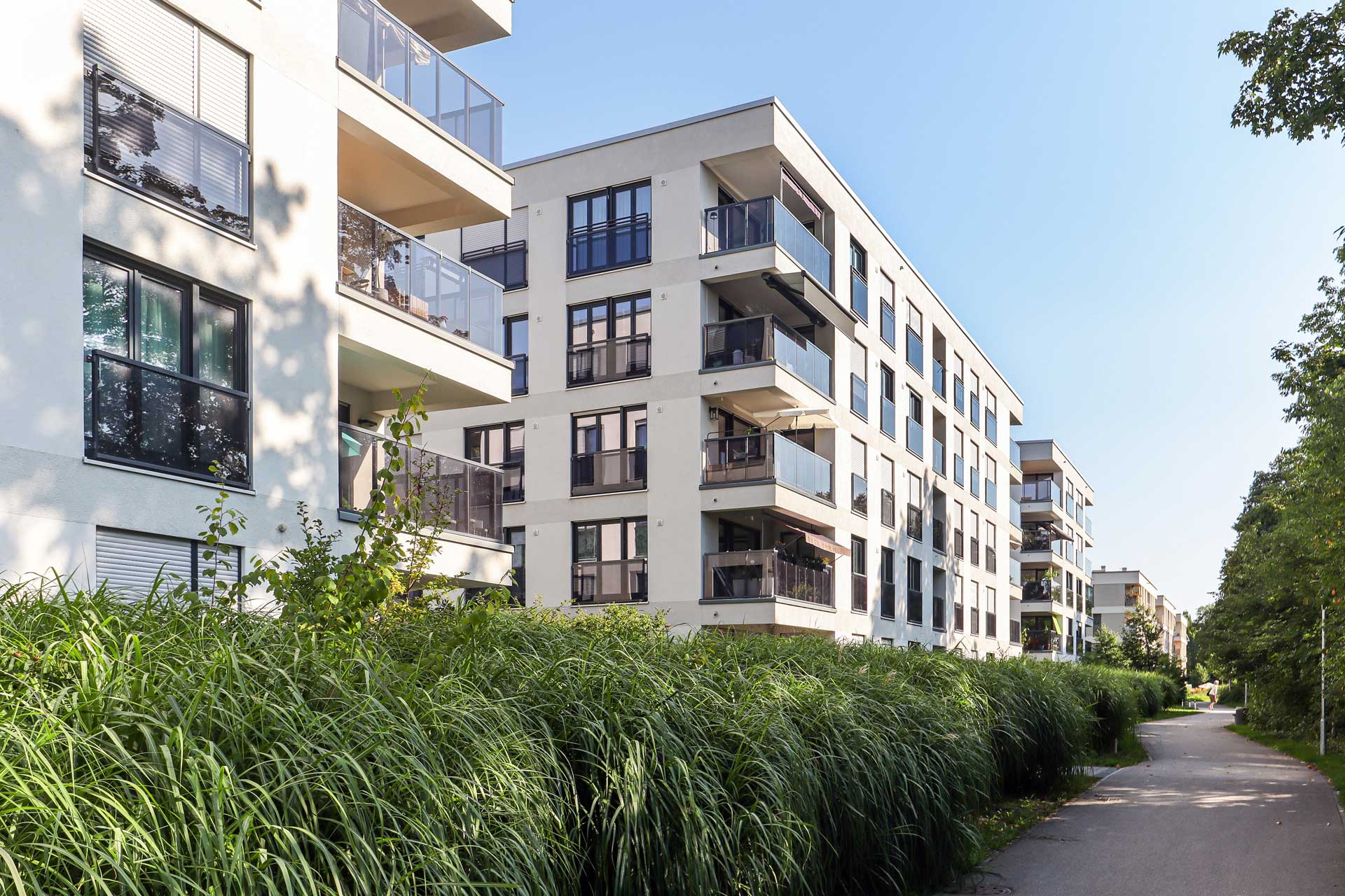 Mehrfamilienhäuser mit Balkonen an einem grünen Weg und hohem Gras.