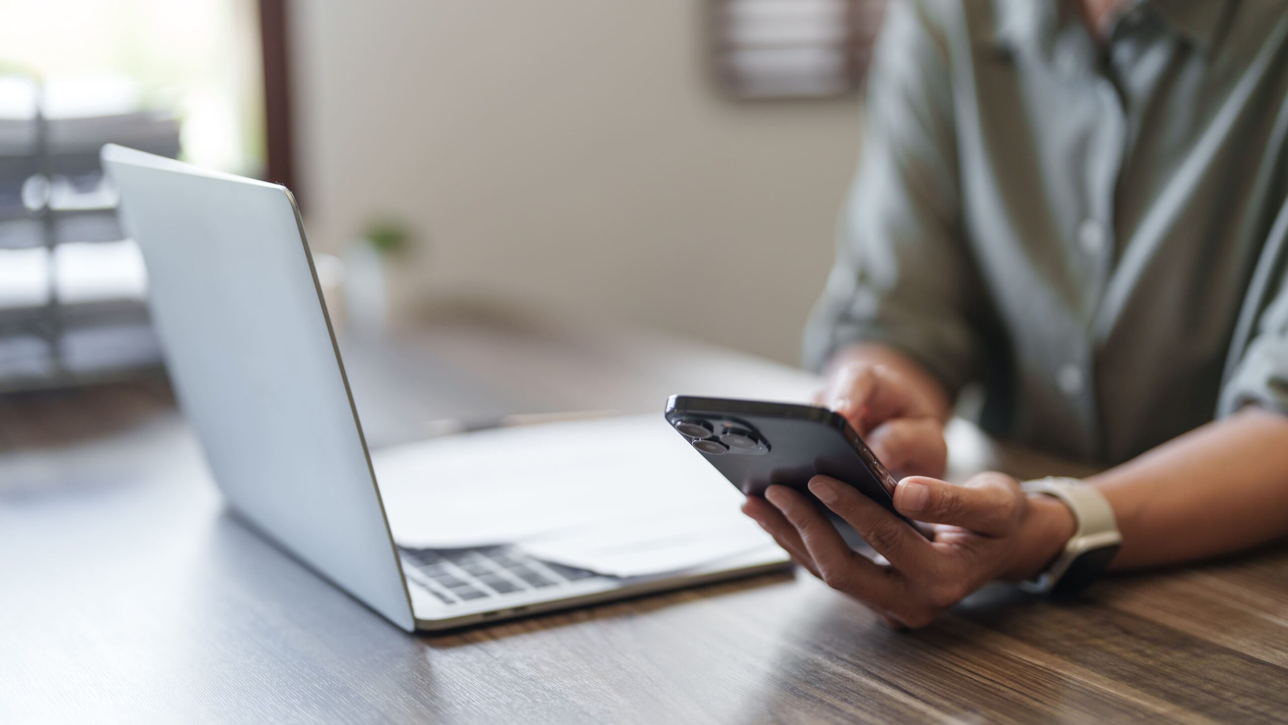Person am Schreibtisch mit Smartphone, Laptop und Papieren.