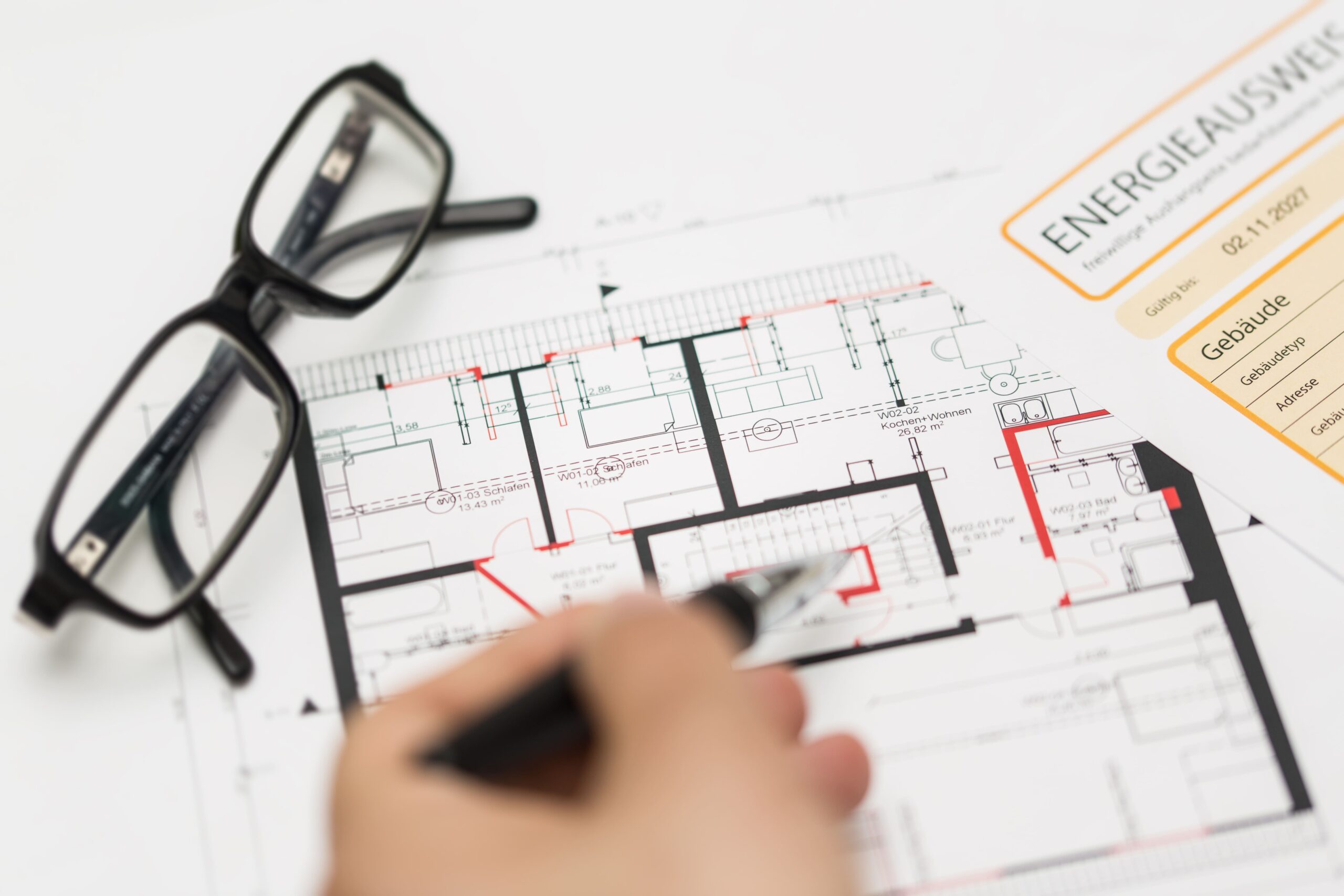 Hand mit Stift zeigt auf Gebäudeplan neben Brille und Zertifikaten.