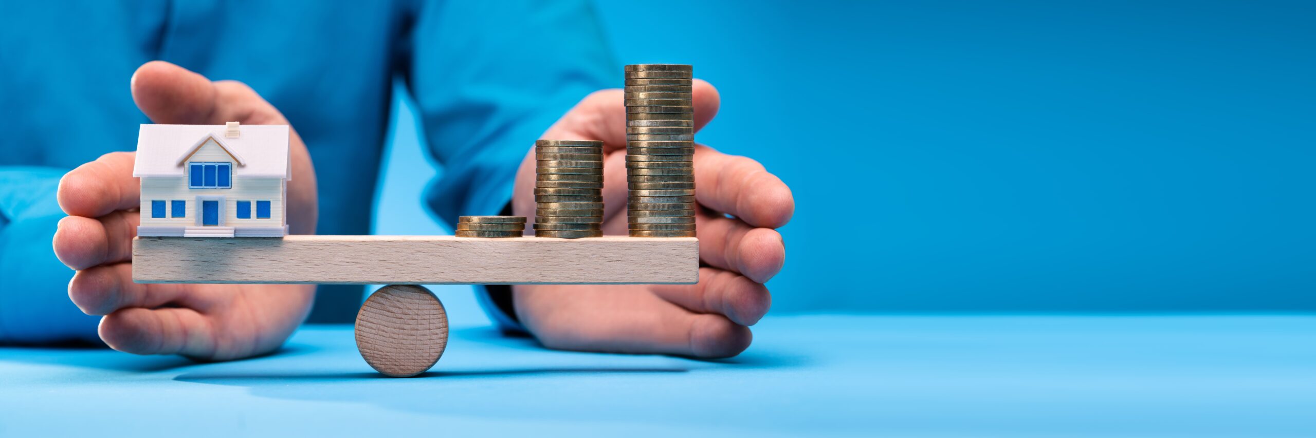 Modellhaus und Münzen auf Holzplanke vor blauem Hintergrund.
