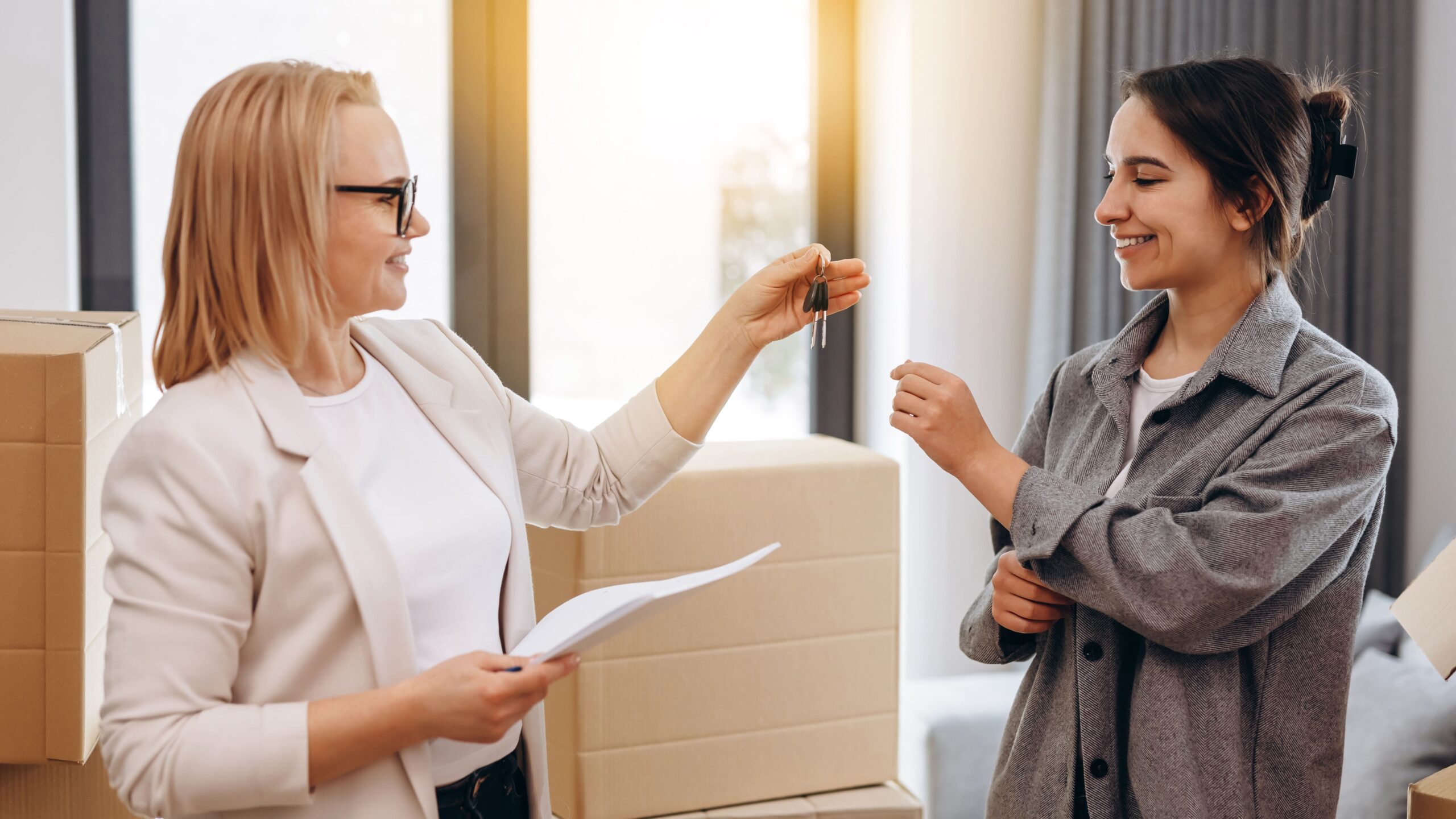 Frauen übergeben Schlüssel, lächeln, Umzugskartons im Hintergrund.