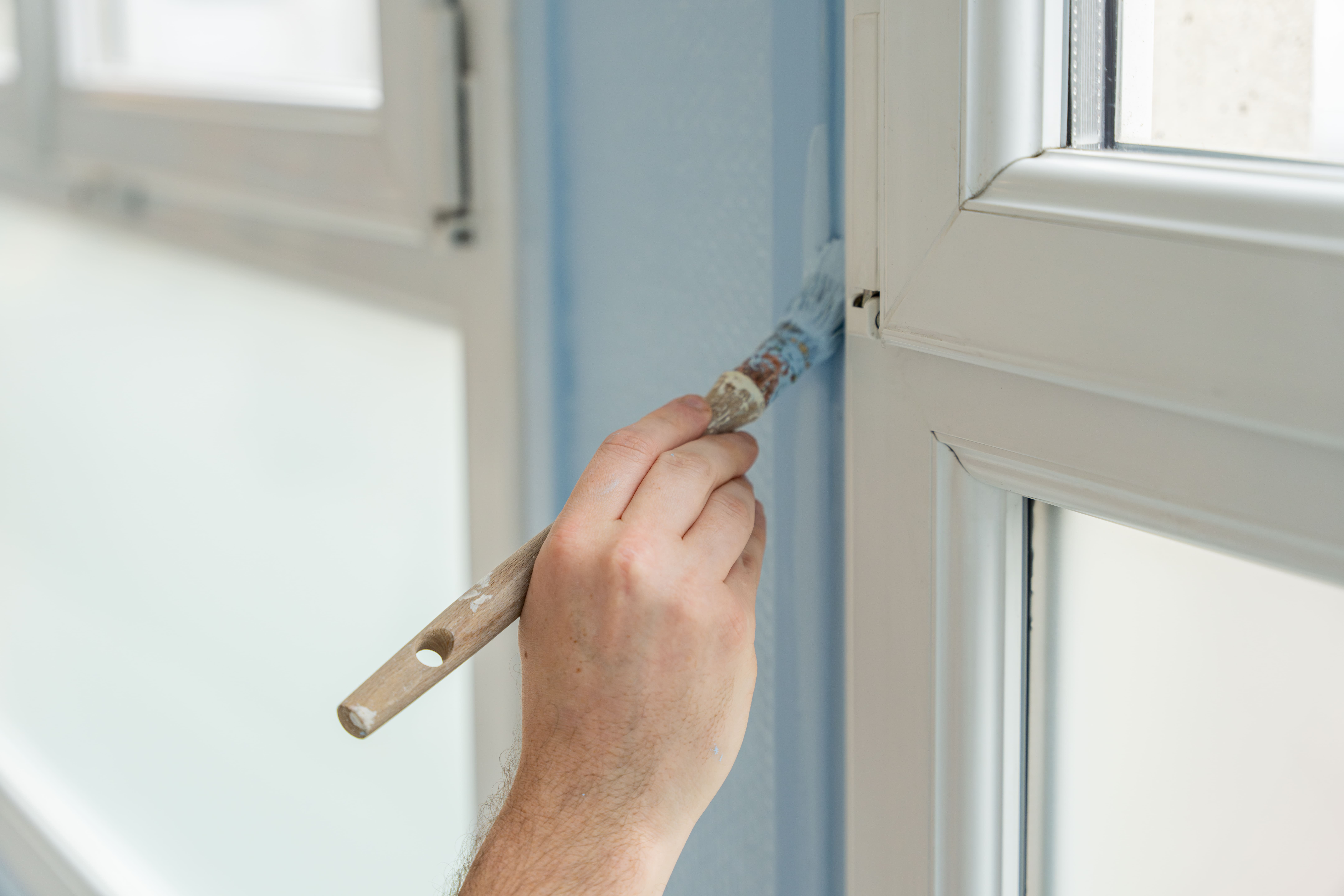 Hand streicht blaue Wand mit Pinsel neben weißem Fensterrahmen.
