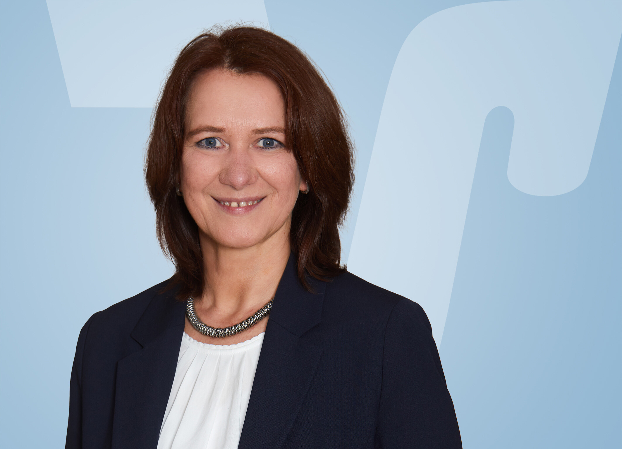 Frau mit braunen Haaren, Navy Blazer, lächelt vor blauem Hintergrund.