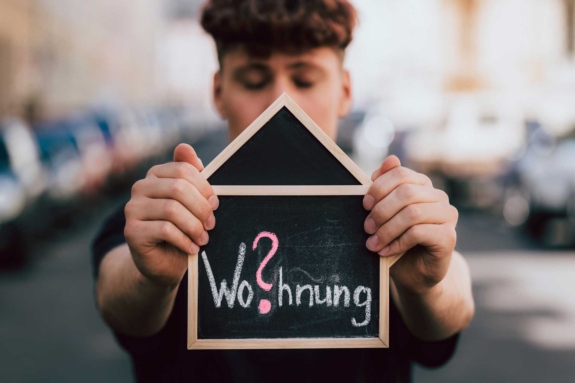 Junger Mann hält Tafel mit Aufschrift 'Wohnung?' in den Händen: Wohnraummangel Berlin