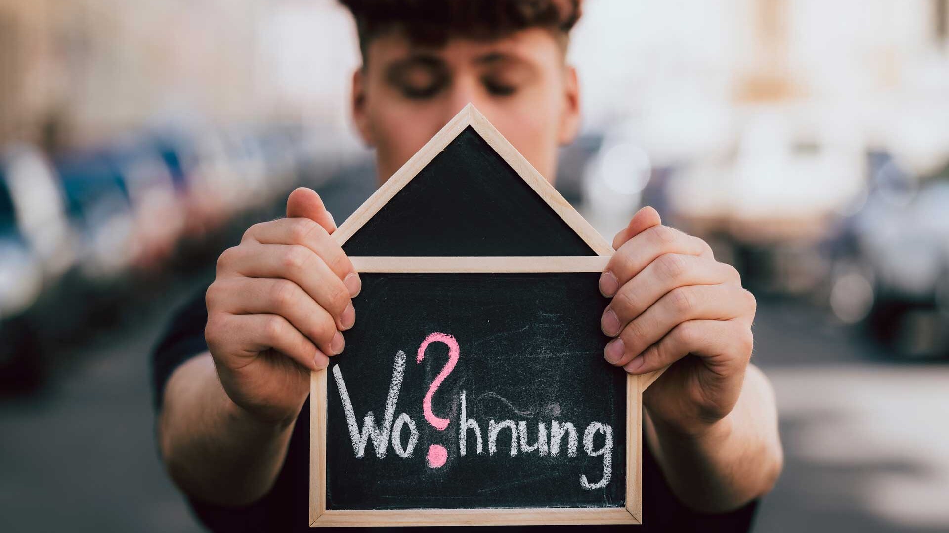 Junger Mann hält Tafel mit Aufschrift 'Wohnung?' in den Händen: Wohnraummangel Berlin