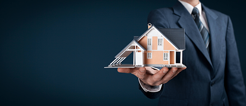 Person im Anzug hält ein Modellhaus vor dunklem Hintergrund.