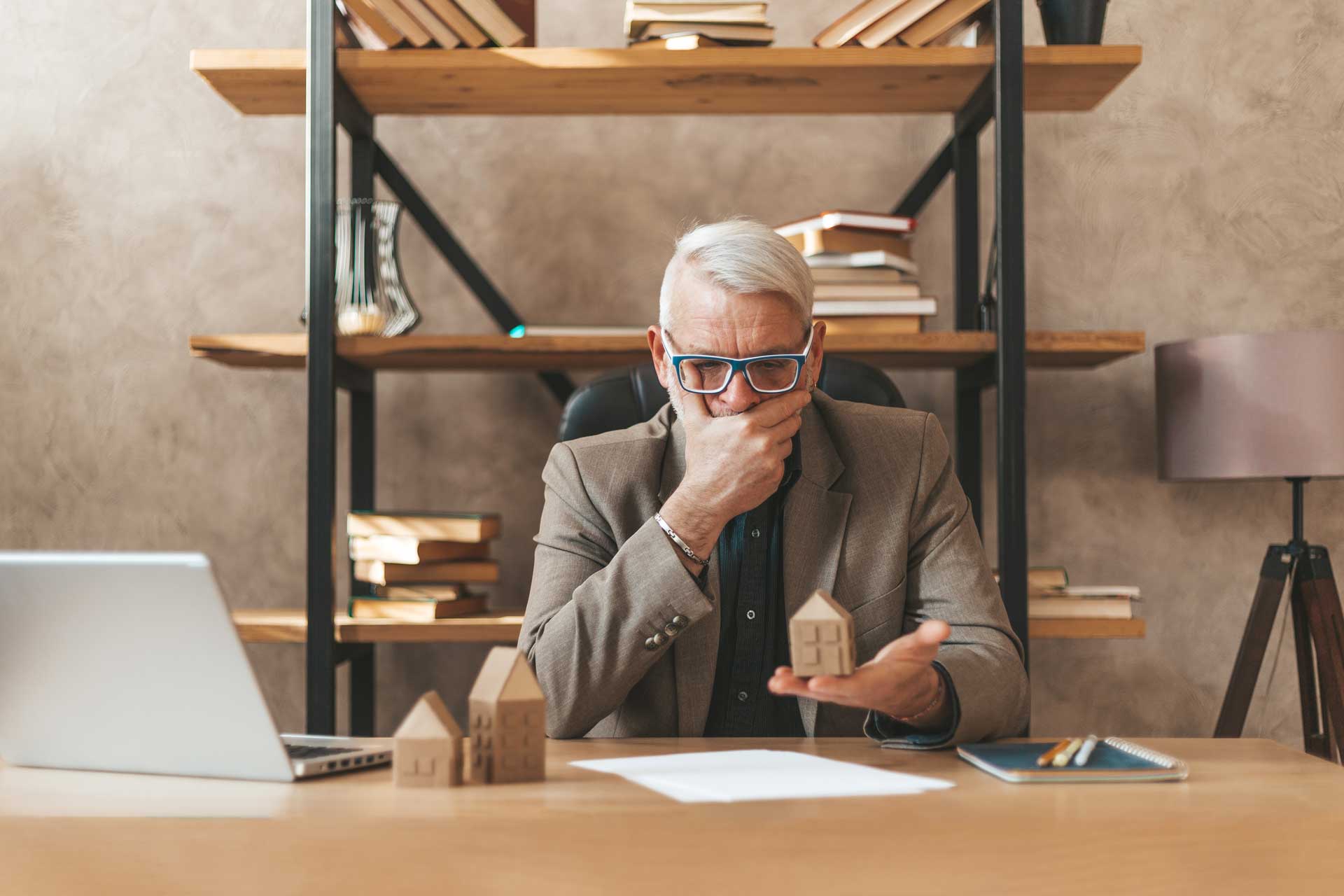 Mann am Schreibtisch mit Modellhaus überlegt, ob er privat oder über Makler verkaufen soll