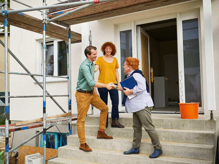 Ein Paar begrüßt die Maklerin vor einem Haus, das gerade noch eine Baustelle ist.