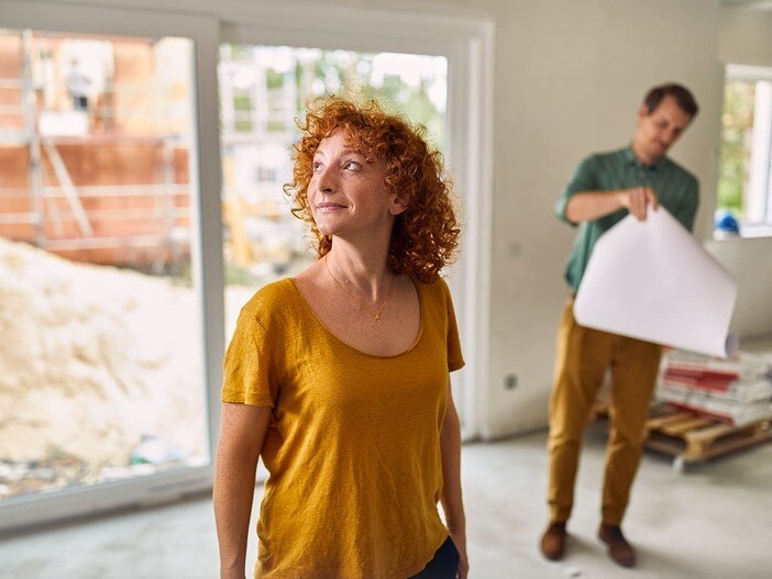Frau steht im Rohbau eines Hauses und sieht sich um, im Hintergrund steht ein Mann mit Bauplänen