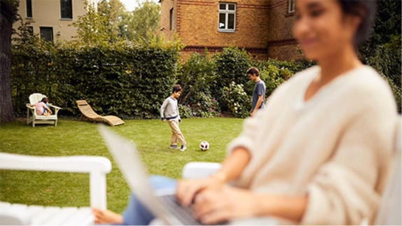 Frau am Laptop im Garten, Kinder spielen Fußball.