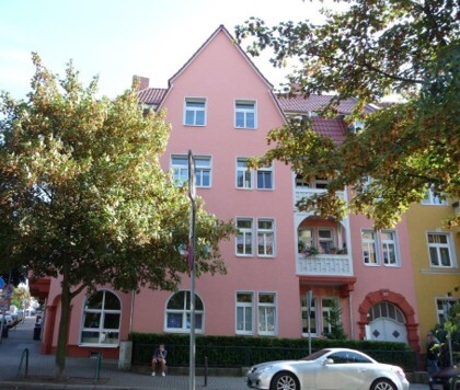 Pinkes Gebäude mit Bögen, Bäumen und silbernem Auto davor.