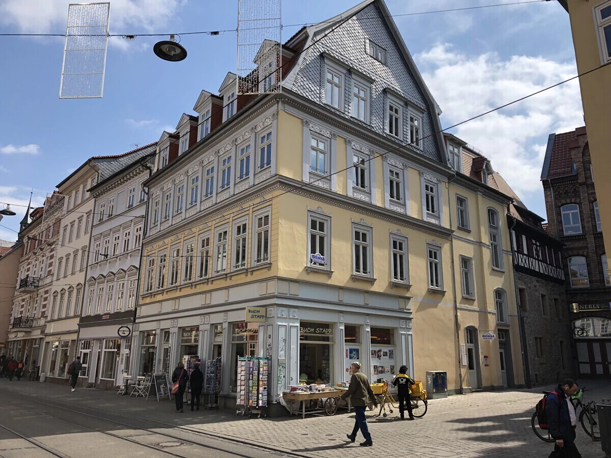 Eckhaus mit Geschäften, Passanten und blauem Himmel mit Wolken.