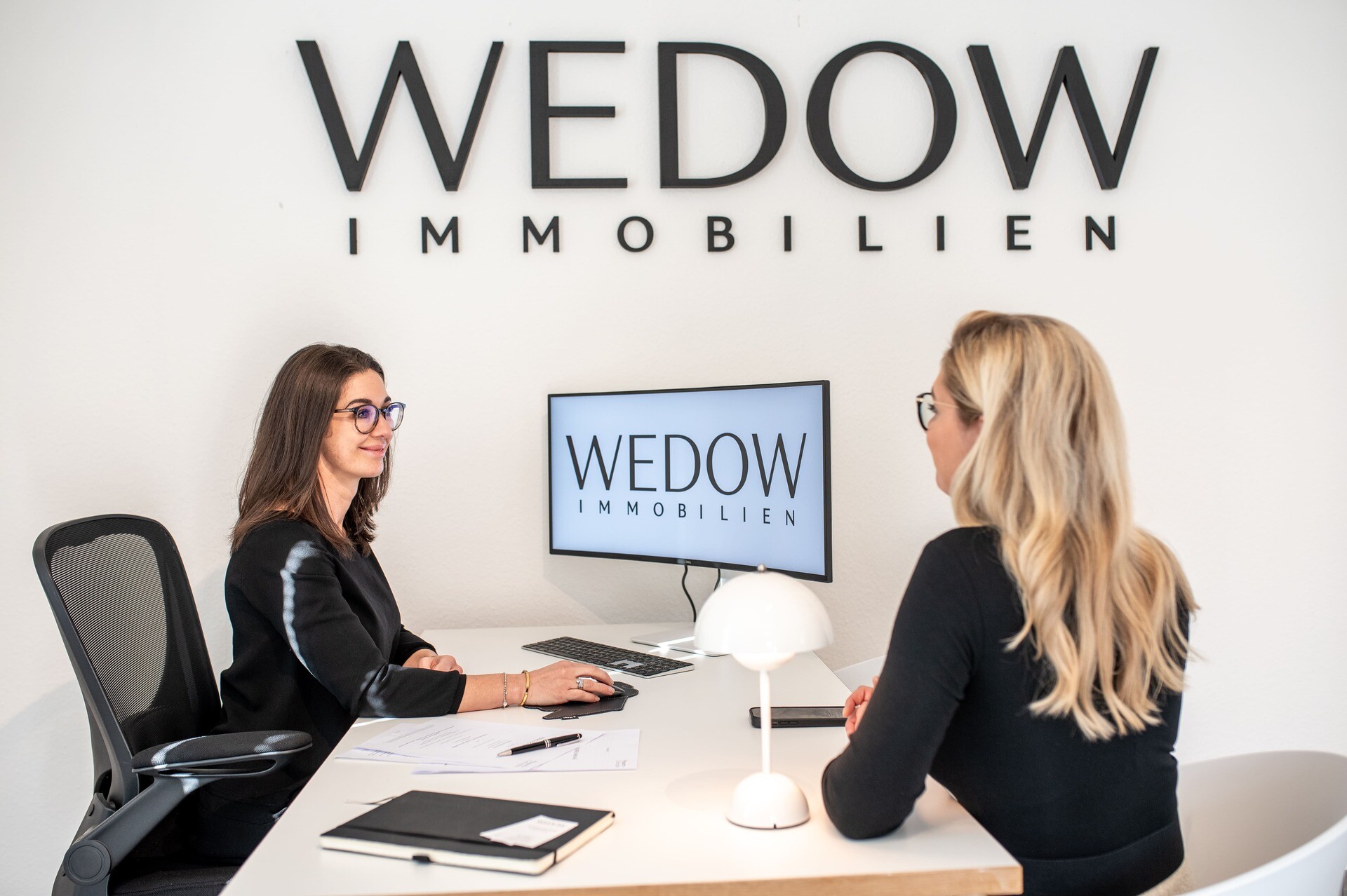 Zwei Frauen am Schreibtisch, WEDOW IMMOBILIEN Schilder im Büro.