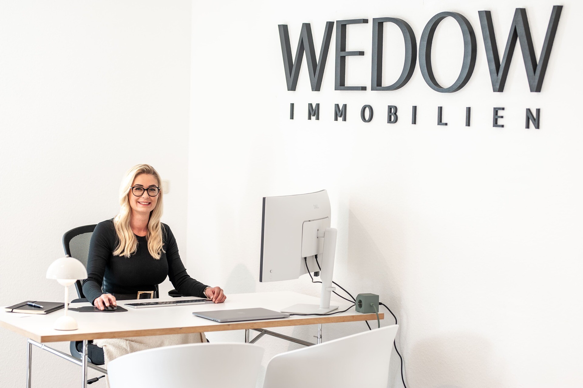 Frau lächelt am Schreibtisch vor WEDOW IMMOBILIEN-Schriftzug.