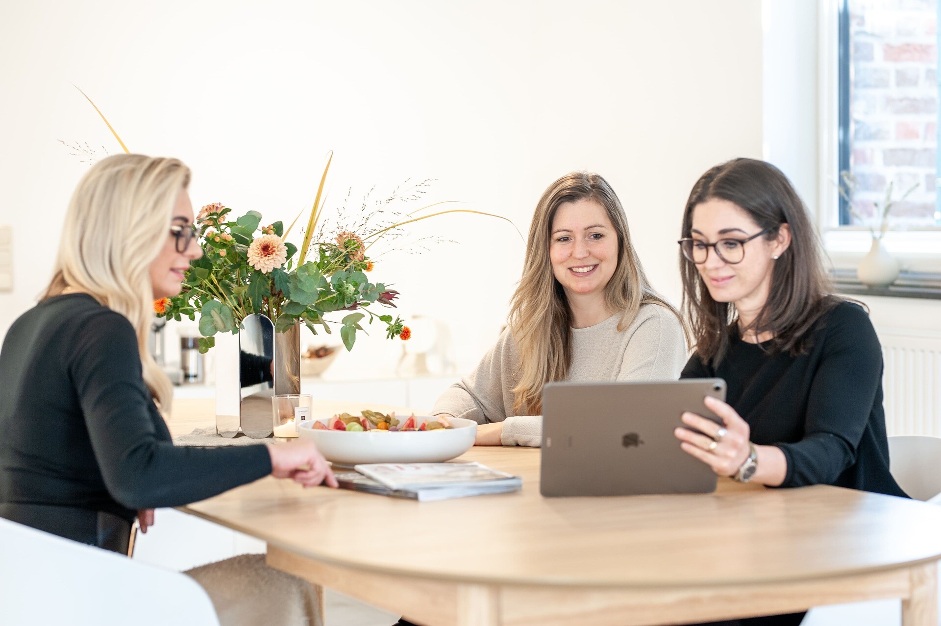 Drei Frauen lächeln am Tisch und schauen auf ein Tablet.