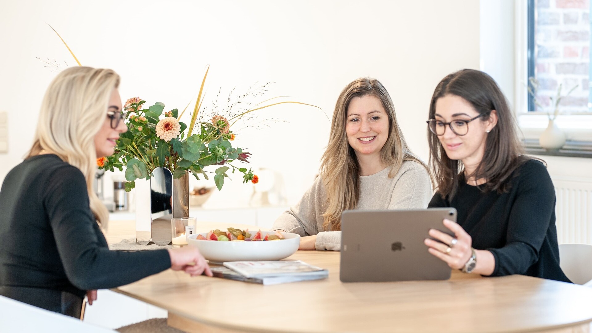 Drei Frauen lächeln am Tisch und schauen auf ein Tablet.