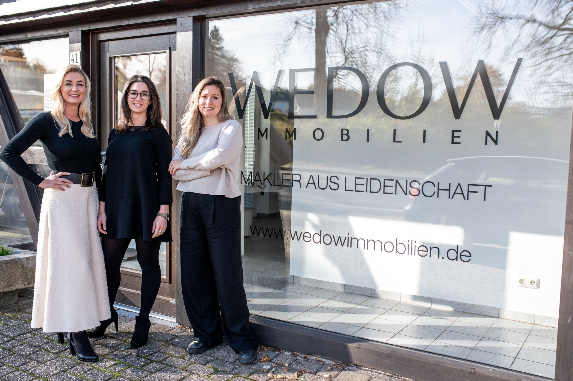 Drei lächelnde Frauen vor Glastür mit WEDOW IMMOBILIEN-Schild.