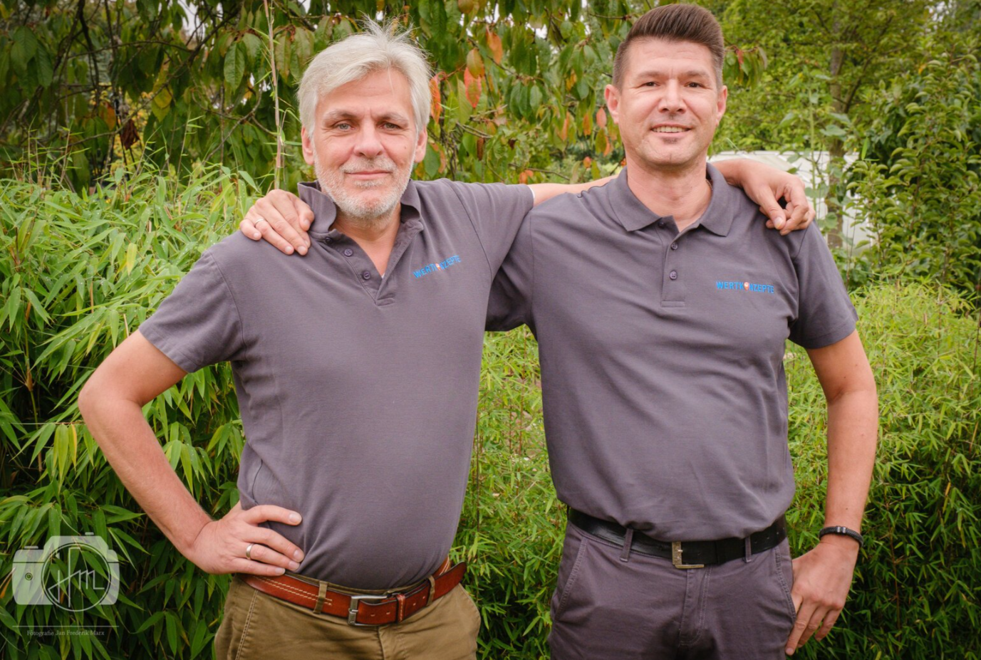 Zwei Männer in grauen Poloshirts umarmen sich draußen und lächeln.