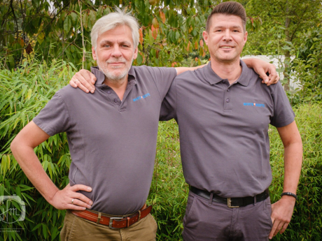 Zwei Männer in grauen Poloshirts umarmen sich draußen und lächeln.