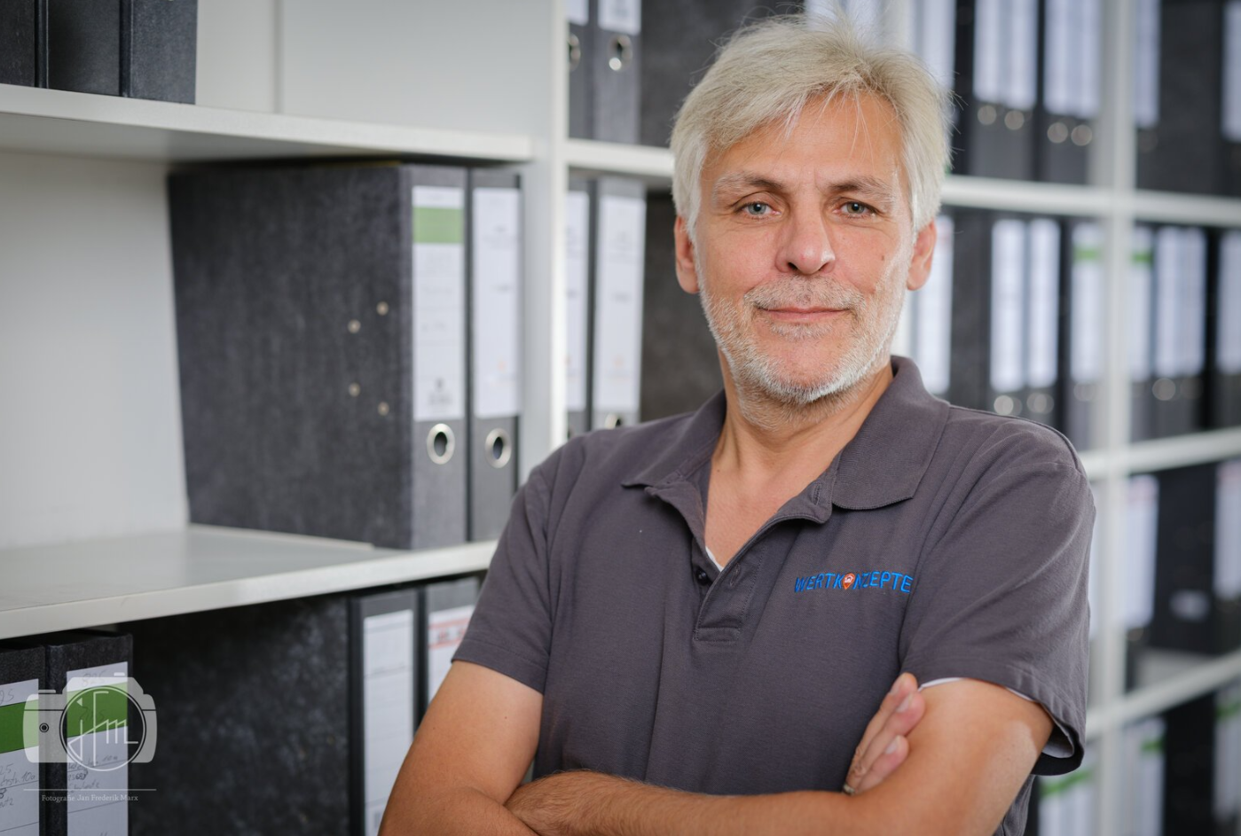 Älterer Mann mit grauem Bart lächelt, Arme vor Büroregalen verschränkt.