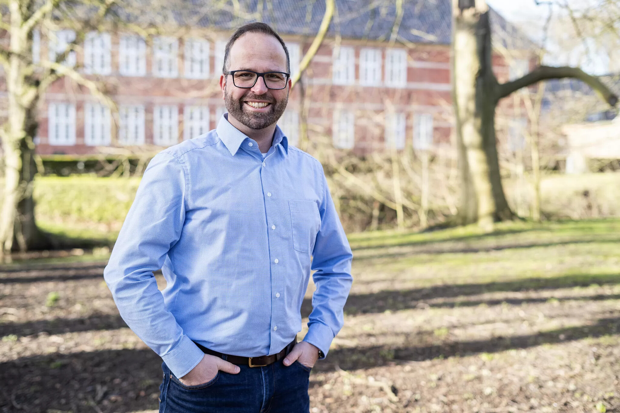 Mann im blauen Hemd steht draußen im sonnigen Park vor Bäumen.