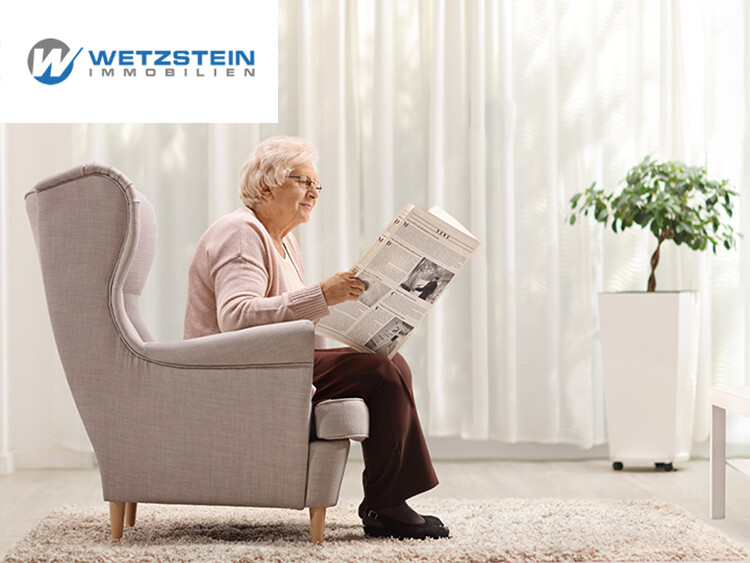 Ältere Person sitzt in einem grauen Ohrensessel und liest eine Zeitung in einem hellen Raum mit weißen Vorhängen und einem grünen Pflanzenkübel