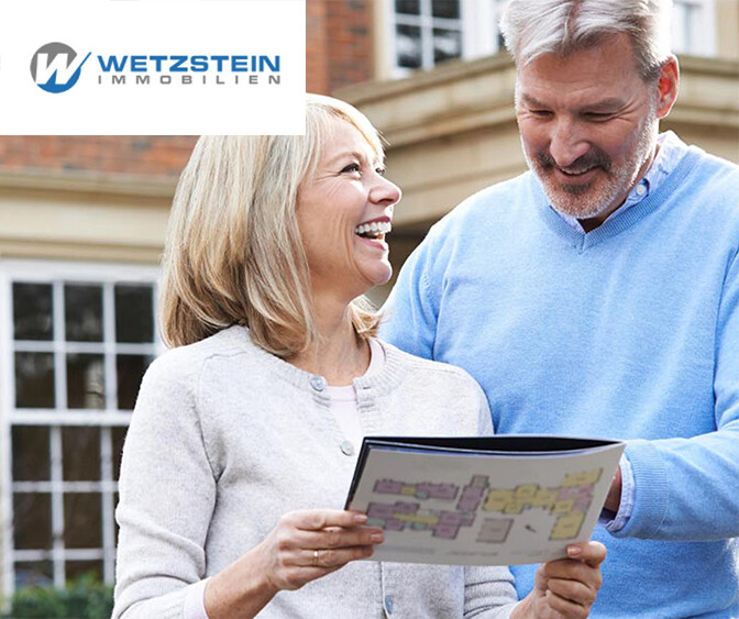 Älteres Paar steht vor einem Backsteinhaus und betrachtet gemeinsam einen Grundrissplan