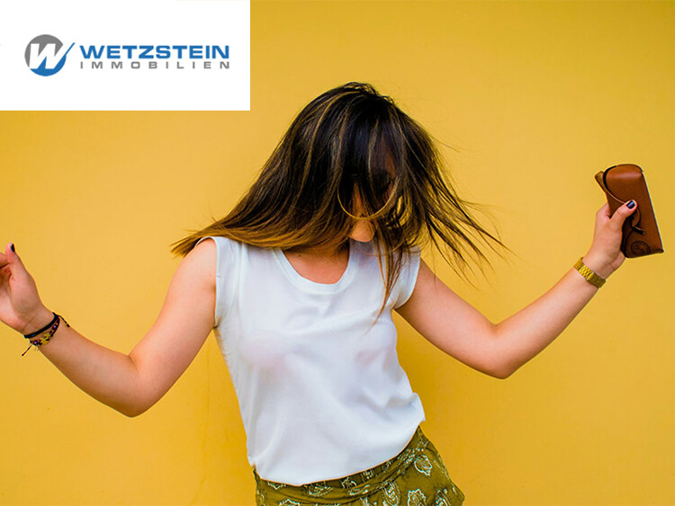 Frau mit langen braunen Haaren tanzt vor gelber Wand, trägt weiße Bluse und grüne Hose, hält braunes Portemonnaie