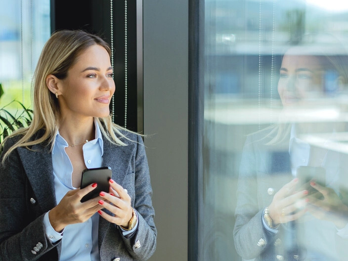 Geschäftsfrau mit Smartphone blickt aus dem Fenster.